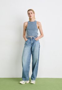 Personne avec les cheveux blonds courts portant un crop top sans manches bleu, un jean bleu ample et des baskets blanches se tenant contre un mur blanc.