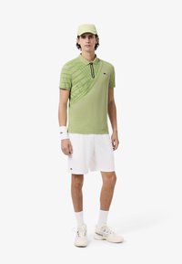 Polo shirt verde claro con un patrón geométrico, cremallera negra, combinado con pantalones cortos blancos, pulsera blanca y zapatillas beige de Lacoste.