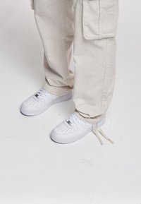 Hvite skinn-sneakers med perforert tåboks, flate lisser og en tykk gummisåle, paret med beige kordfløyelsbukser med sidelommer.