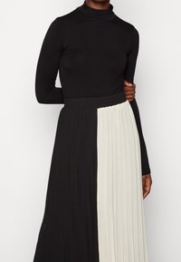 Top a maniche lunghe e collo alto di colore nero, abbinato a una gonna plissettata con un design a blocchi di colore in nero e crema. Tessuto morbido e elasticizzato.
