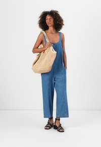 Salopette in denim blu, design senza maniche con gambe ampie e tasche laterali, abbinata a una borsa intrecciata beige testurizzata e sandali neri.
