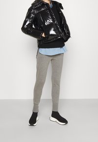 Skinnende sort pufferjakke, cropped design med hætte, båret over en sort sweater og en lyseblå skjorte, parret med grå leggings og sorte sko.