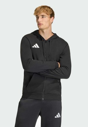 Junger Mann mit hellbraunen Haaren, trägt eine schwarze Adidas Kapuzenjacke mit Reißverschluss und passende schwarze Hose mit weißen Adidas-Logos, Arme verschränkt, schaut nach links.