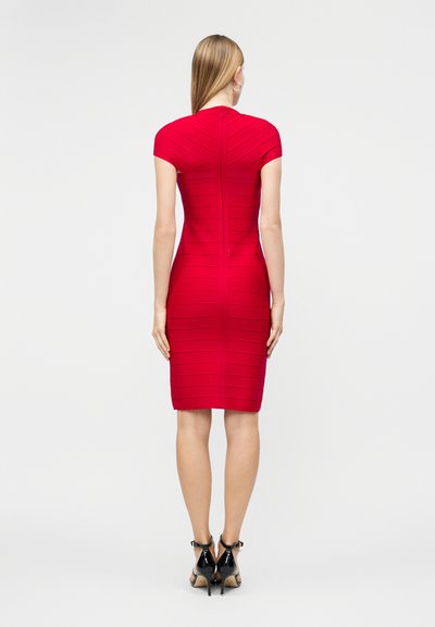 Žena s dlouhými rovnými vlasy stojí zády otočená, oblečená v přiléhavých červených šatech do kolen a černých vysokých podpatcích na jednobarevném pozadí.