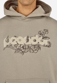 Beżowa bluza z kapturem z dużą teksturowaną grafiką kwiatową i jasnym logo marki z przodu. Miękki materiał, luźny krój.