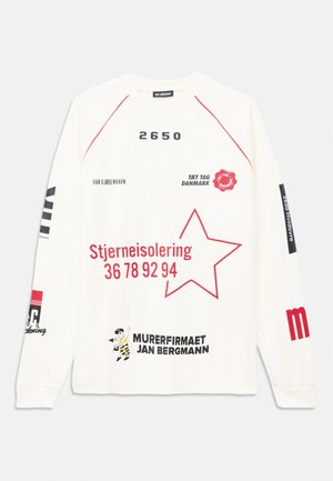 Balta garām piedurknēm krekls ar sarkano un melno tekstu, sarkano zvaigznes kontūru un grafiskajiem logotipiem, kas izvietoti uz priekšpuses un piedurknēm.