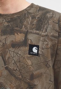 Κοντινή λήψη ενός πουκαμίσου Carhartt με σχέδιο παραλλαγής, που διαθέτει καφέ και πράσινο σχέδιο φύλλων και κλαδιών με μαύρο τετράγωνο λογότυπο Carhartt.