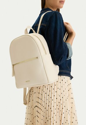 Žena nosící krémový kožený batoh s přední kapsou na zip, v kombinaci s tmavou džínovou bundou a krémovou plisovanou sukní s puntíky.