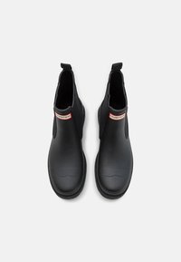 Bottines noires en caoutchouc lisse avec des panneaux latéraux en élastique, un bout arrondi et une languette ; présente une étiquette de marque rouge et blanche.