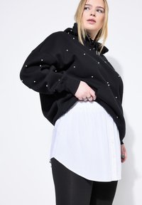 Sudadera negra con adornos de perlas dispersos, superpuesta a una camisa blanca con botones. Leggings negros completan el conjunto.