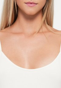 Bijela sleeveless majica s širokim izrezom. Mekani, priljubljeni materijal koji ističe ramena i ključnu kost. Glatki, minimalistički dizajn.