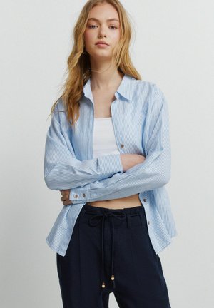 Młoda kobieta o długich blond włosach, ubrana w jasnoniebieską koszulę w paski zapinaną na guziki, noszoną na białym krótki topie, oraz czarne spodnie ze sznurkiem w pasie.