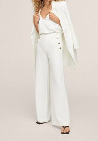 Witte op maat gemaakte outfit met wijde broekspijpen en gouden knopen, een v-hals top en een bijpassende blazer, wat zorgt voor een samenhangende, elegante uitstraling.