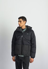 Black puffer jacket with a hood, zippered front, and elastic cuffs. Features a block pattern and a logo patch on the chest.