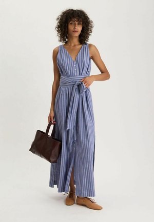 Femme aux cheveux bouclés portant une robe maxi rayée bleu et blanc sans manches, tenant un sac à main marron foncé et portant des chaussures couleur caramel.