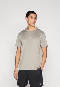 Αθλητική μπλούζα σε ανοιχτό γκρι χρώμα από λείο ύφασμα, με στρογγυλή λαιμόκοψη και κοντά μανίκια, με ένα μικρό λευκό λογότυπο στο στήθος.