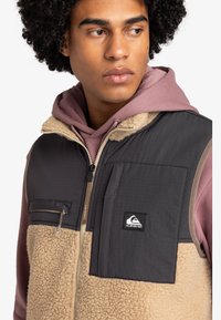 Weste aus dunkelgrauem Nylon und beigem Fleece. Verfügt über einen Reißverschluss vorne, eine Brusttasche und ein Logo-Patch. Über einem pinken Hoodie getragen.