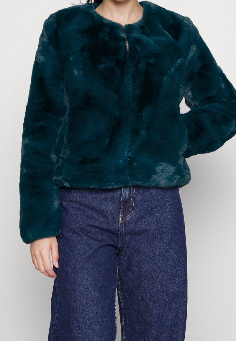 Veste en fausse fourrure teal avec un col rond, des manches longues et une coupe courte. Texture douce et sans fermetures visibles. Associée à un pantalon en denim.