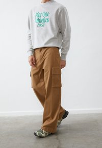 Sudadera gris con "Pier One Athletics 1968" en texto verde, combinada con pantalones cargo marrones con bolsillos laterales y zapatillas verdes.