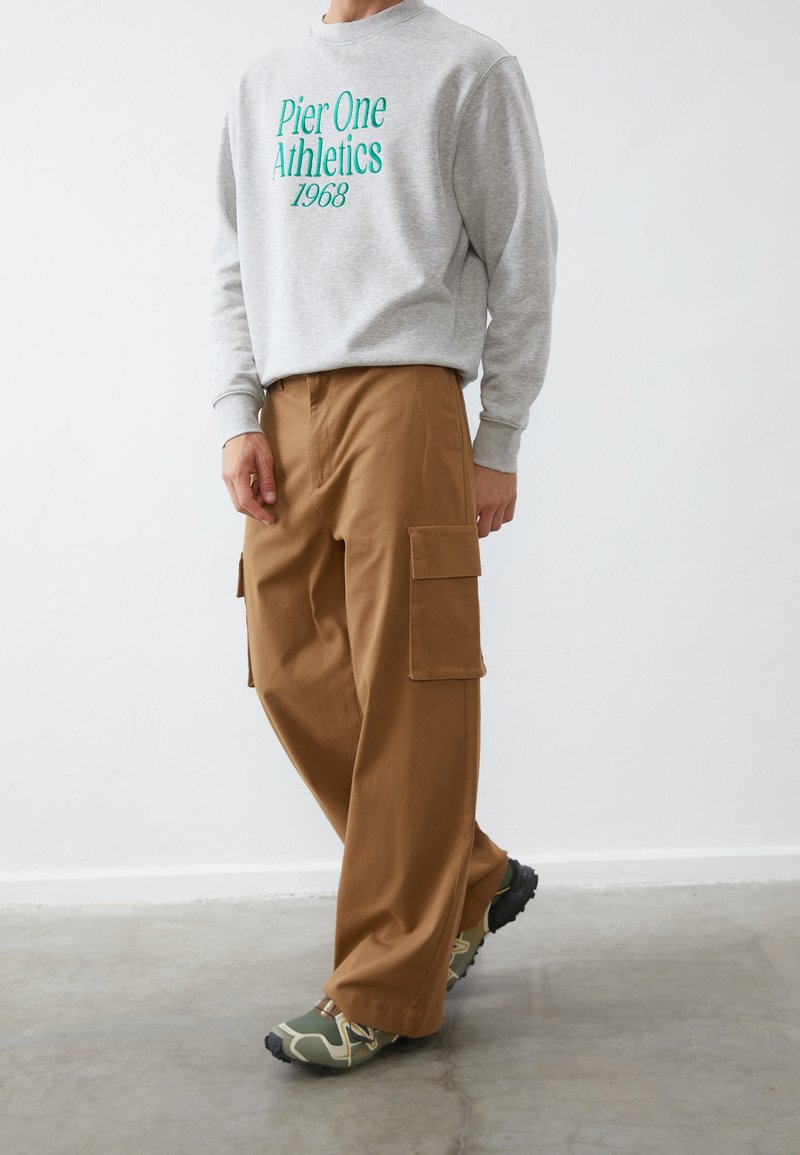 Sudadera gris con "Pier One Athletics 1968" en texto verde, combinada con pantalones cargo marrones con bolsillos laterales y zapatillas verdes.
