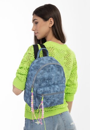 Zaino blu denim con un motivo trapuntato, dotato di tasca frontale e accenti di cordoncino colorati, posizionato su un maglione verde brillante.