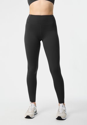 Osoba nosiace čierne legíny s vysokým pásom a svetlé tenisky, stojaca pred jednoliatym bielym pozadím.