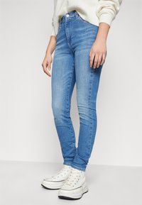 Jeans ajustados de mezclilla en azul claro. Presentan una cintura de mediana altura, diseño de cinco bolsillos y un ligero desgaste en los muslos. Combinados con zapatillas blancas.