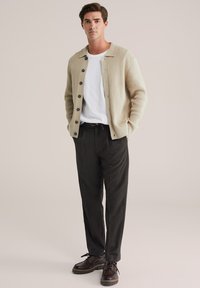 Cardigan bej tricotat, cu guler și nasturi negri, purtat peste un tricou alb, asortat cu pantaloni închisi la culoare și pantofi maro.