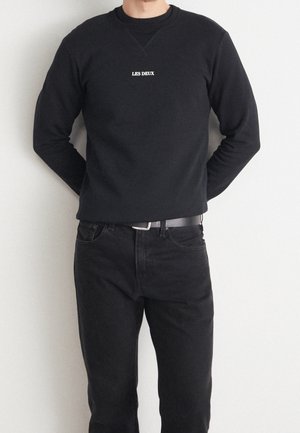 Uomo con felpa nera a maniche lunghe con la scritta "LES DEUX", jeans neri e cintura nera, in piedi contro una parete bianca.