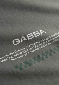 Tissu gris avec le texte blanc "GABBA" et un paragraphe, accompagné en dessous du texte d'un motif à carreaux vert.
