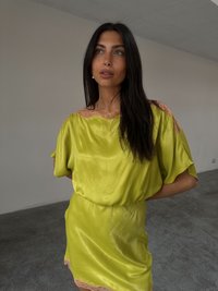 En ung kvinne med langt mørkt hår står mot en nøytral bakgrunn, iført en lys gul kjole med blonde detaljer. Hun ser tankefull ut.