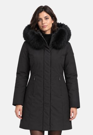 Donna che indossa un cappotto invernale nero di media lunghezza con un grande cappuccio nero rifinito in pelliccia e tasche anteriori con cerniera, in piedi davanti a uno sfondo semplice.