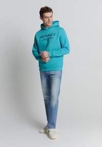 Turquoise hoodie met grafische tekst op de voorkant, geribbelde boorden en zoom, gecombineerd met lichtblauwe jeans en witte sneakers. Eenvoudige achtergrond.