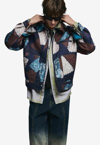 Kolorowa kurtka dżinsowa w stylu patchwork, z niebieskimi, brązowymi i białymi wzorami, z kieszenią na zamek i luźnym krojem. Nosi się ją na jasnej koszuli.