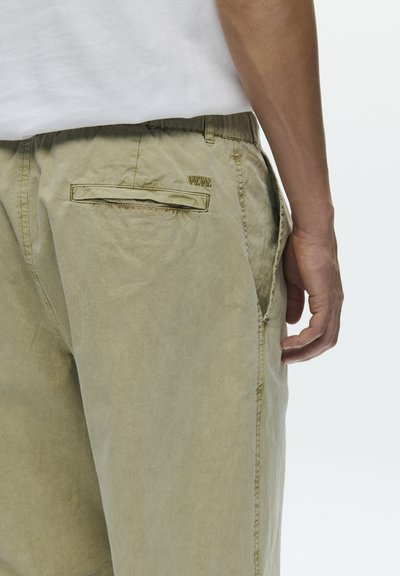 Férfi, aki bézs színű nadrágot visel hátsó zsebbel és "W.W." hímzéssel, valamint fehér inget, egy egyszerű világos háttér előtt áll.