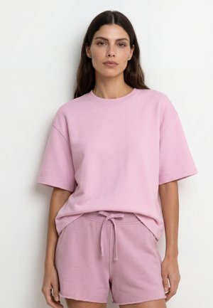 Vrouw draagt een loszittend pastelroze T-shirt met korte mouwen en bijpassende short met trekkoord, staand tegen een effen lichte achtergrond.