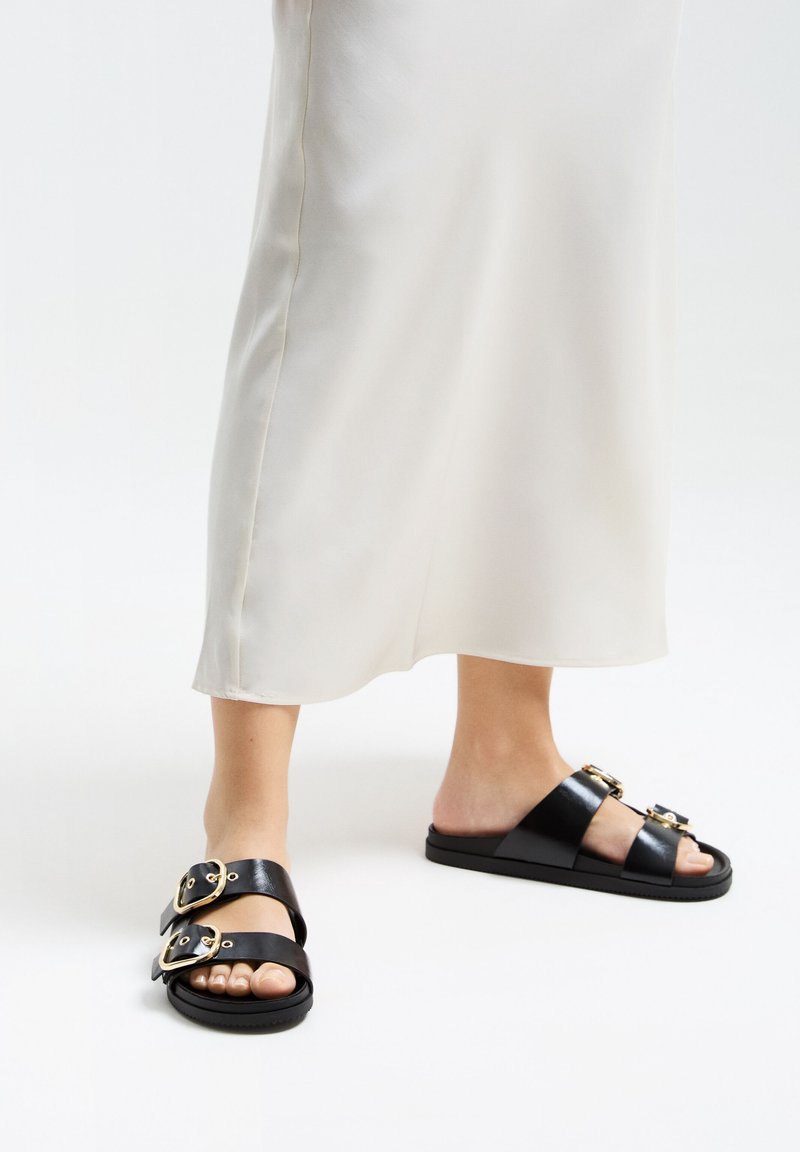Black leather sandals with two wide straps featuring gold buckles. Thick sole and smooth texture complete the design.
