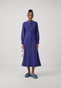 Abito di lino viola con lunghe maniche a sbuffo, chiusura con bottoni e dettagli ricamati. Abbinato a sandali blu a scivolo e una borsa rosa.