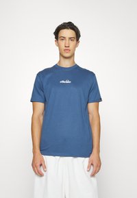 Blå bomulds T-shirt med rund hals og korte ærmer, der har et hvidt logo grafisk design på brystet. Modellen har lyse shorts på.