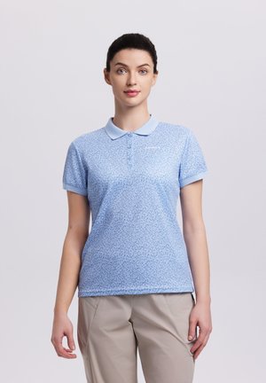 Femme aux cheveux foncés portant un polo bleu clair à motifs et un pantalon beige, debout devant un fond uni clair.