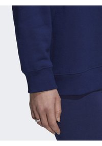 Ruka s stříbrným prstenem odpočívající vedle osoby oblečené v tmavě modrém dlouhém rukávu a odpovídajících tmavě modrých kalhotách.