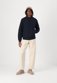 Veste zippée marine avec un col montant, des poignets côtelés et un petit logo. Associée à un pantalon crème clair et des chaussures beiges.