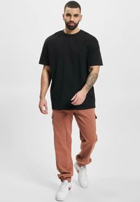 Black cotton t-shirt paired with loose, rust-colored cargo pants featuring side pockets. White sneakers with red accents complete the outfit.