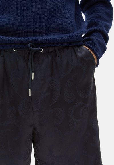 Navyblauer Strickpullover mit gerippter Textur, kombiniert mit dunkel gemusterten Shorts mit Kordelzug, die ein dezentes Wirbelmuster und silberne Hardware-Akzente aufweisen.