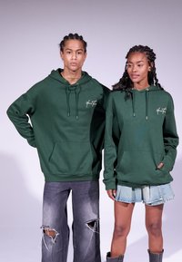 Sweatshirts à capuche verts avec poches avant, cordons de serrage et logo ; portés avec un jean gris usé et une jupe en denim. Environnement de studio.