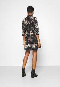 Schwarzes Blumenmusterkleid mit Dreiviertelärmeln, geschichteter Gestaltung und volantartigem Saum. Kombiniert mit Stiefeletten, die einen Blockabsatz haben.