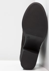 Semelle de chaussure noire avec des motifs de grip texturés et marquage de taille 38, positionnée sur une surface blanche.