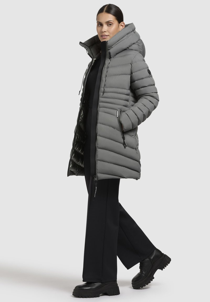 Cappotto puffy grigio con cappuccio, caratterizzato da cuciture orizzontali, tasche con zip e finitura resistente all'acqua, indossato sopra un outfit nero.