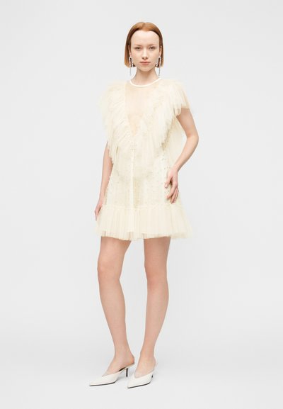 Φόρεμα από ελεφαντόδοντο τούλι με στρώσεις βολάν, βαθύ V-λαιμόκοψη και υφασματικές λεπτομέρειες. Συνδυασμένο με λευκά μυτερά mules και μεγάλα σκουλαρίκια.