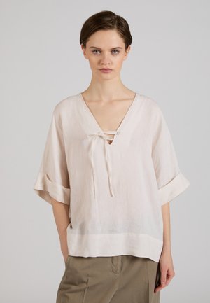 Femme aux cheveux courts portant un chemisier ample beige clair à col en V avec une fermeture à nouer et des manches mi-longues retroussées, associé à un pantalon kaki.